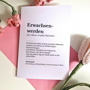 May include: A white card with the German word "Erwachsenwerden" and its definition, resting on a pink envelope. Crochet pink flowers and stems are arranged around the card, creating a soft, handmade aesthetic.