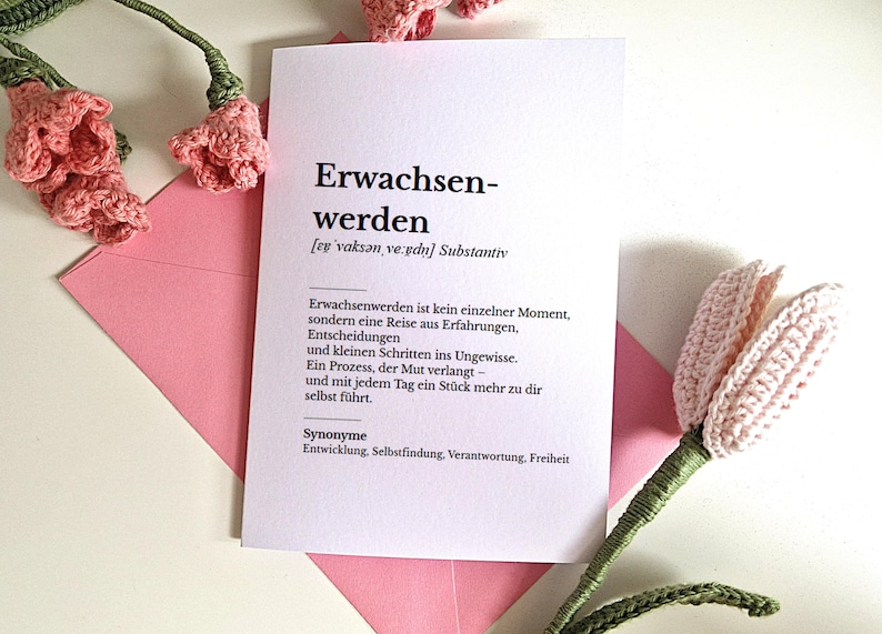 May include: A white card with the German word "Erwachsenwerden" and its definition, resting on a pink envelope. Crochet pink flowers and stems are arranged around the card, creating a soft, handmade aesthetic.
