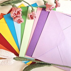 May include: A collection of colorful envelopes fanned out, showcasing a spectrum of hues from red and orange to yellow, blue, green, and purple. Crochet flowers and stems are artfully arranged around the envelopes, adding a handcrafted touch.
