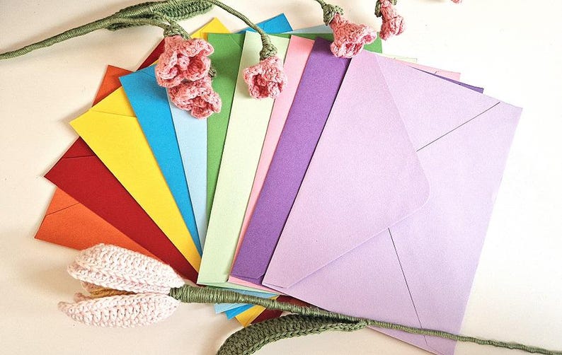 May include: A collection of colorful envelopes fanned out, showcasing a spectrum of hues from red and orange to yellow, blue, green, and purple. Crochet flowers and stems are artfully arranged around the envelopes, adding a handcrafted touch.