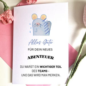 May include: A white card with German text and a cartoon mouse illustration. The card is placed on a pink envelope, with pink crocheted flowers. The text reads "Alles Gute", "FÜR DEIN NEUES ABENTEUER", and other phrases.
