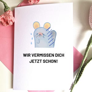May include: A white card featuring a watercolour illustration of a crying mouse, with the German text "WIR VERMISSEN DICH JETZT SCHON!". The card is placed in a pink envelope, with pink crocheted flowers in the background.