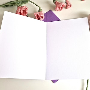 May include: An open, blank greeting card with a purple envelope peeking out from the bottom. Crochet pink flowers and green stems are arranged around the card, creating a floral theme. The card is white.