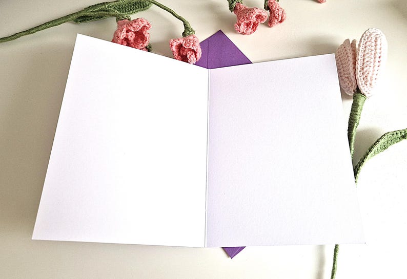 May include: An open, blank greeting card with a purple envelope peeking out from the bottom. Crochet pink flowers and green stems are arranged around the card, creating a floral theme. The card is white.