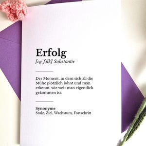 Puede incluir: Una tarjeta blanca con la palabra alemana "Erfolg" impresa en negro, que significa "Éxito". La tarjeta está en un sobre morado, con flores rosas de ganchillo en el fondo.