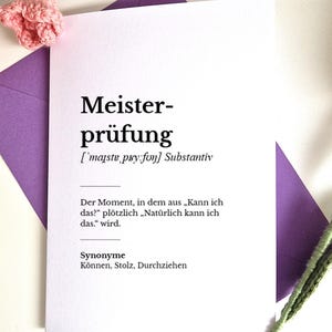 Puede incluir: Una tarjeta blanca con la palabra alemana "Meisterprüfung" impresa en negro, junto con una definición y sinónimos. La tarjeta está colocada sobre un sobre morado y decorada con flores de ganchillo rosas.