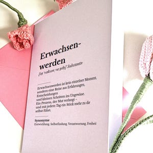 May include: A light pink card with the German word "Erwachsenwerden" printed on it, meaning "growing up". The card is accompanied by pink and green crocheted flowers.