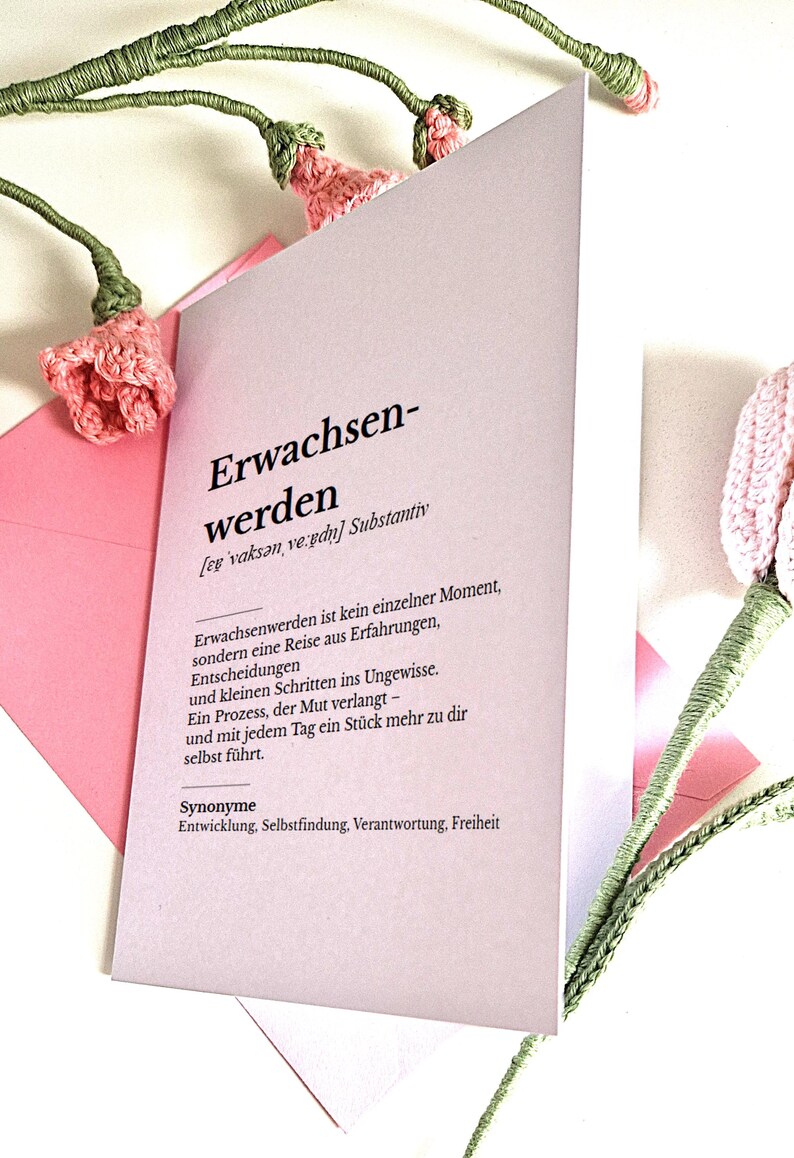 May include: A light pink card with the German word "Erwachsenwerden" printed on it, meaning "growing up". The card is accompanied by pink and green crocheted flowers.