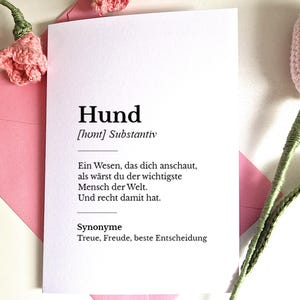 May include: A white card with the German word "Hund" (dog) and its definition, resting on a pink envelope. The card is decorated with pink crocheted flowers and green stems.