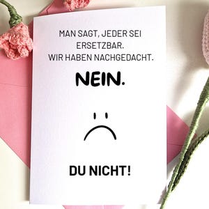 Op de afbeelding: Een witte wenskaart met Duitse tekst en een illustratie van een verdrietig gezicht. De kaart zit in een roze envelop, met roze gehaakte bloemen op de achtergrond. De tekst luidt: "MAN SAGT, JEDER SEI ERSETZBAR. WIR HABEN NACHGEDACHT. NEIN. DU NICHT!"