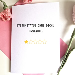 Pode incluir: Um cartão branco com o texto "SYSTEMSTATUS OHNE DICH: UNSTABIL." e uma classificação de uma estrela. O cartão está em um envelope rosa, com flores de crochê rosa no fundo.