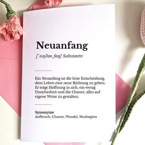 May include: A white card with the German word "Neuanfang" printed in black, meaning "new beginning." The card is in a pink envelope, with pink crocheted flowers in the background.