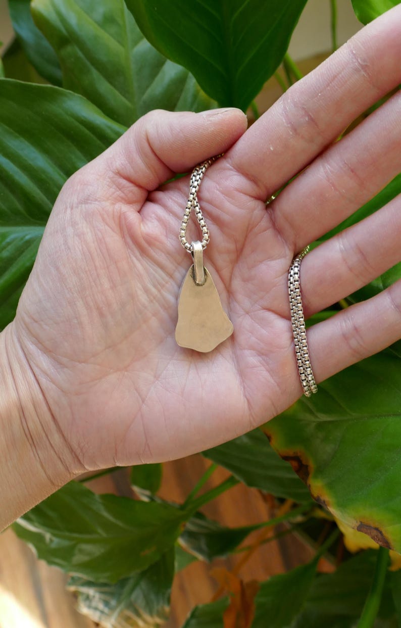 Op de afbeelding: Een zilveren ketting met een gladde, onregelmatig gevormde hanger. De hanger is bevestigd aan een zilveren ketting.