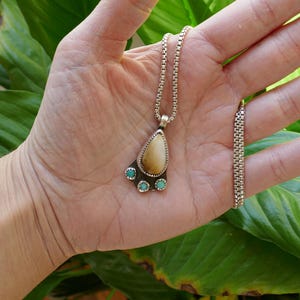 Op de afbeelding: Een zilveren ketting met een hanger met een traanvormig stuk gefossiliseerd ivoor met drie turquoise stenen eromheen.