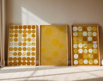 Op de afbeelding: Drie ingelijste abstracte kunstprints. De prints tonen een herhalend patroon van gele en witte cirkels op een witte en gele achtergrond. De frames hebben een licht houten kleur. De kunst wordt tentoongesteld op een beige muur.