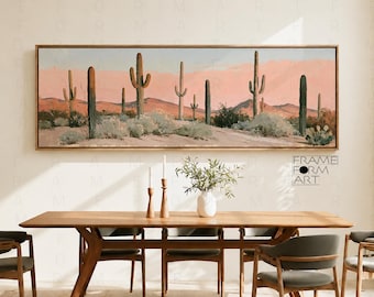 Op de afbeelding: Een horizontale ingelijste wandkunstprint met een woestijnlandschap met hoge saguaro-cactussen, lage woestijnstruiken en verre roze bergen onder een zachte lucht, weergegeven boven een houten eettafel met stoelen en een vaas.