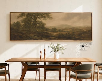 Op de afbeelding: Een panoramisch landschapsschilderij in een dunne houten lijst met een grote boom op de voorgrond, glooiende heuvels en een bewolkte lucht in gedempte aardetinten. Het kunstwerk hangt boven een houten eettafel met stoelen en een vaas.