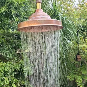 Könnte beinhalten: Eine Außendusche mit Kupferoberfläche. Wasser strömt aus dem Duschkopf, umgeben von üppigem grünem Laub. Der Duschkopf ist an einem Messingrohr befestigt.