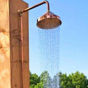 May include: Outdoor copper shower head with water cascading down. The shower head is mounted on a wooden post with copper piping. The shower head is a circular shape with a series of small holes. The background is a clear blue sky and green trees.