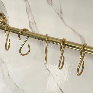 以下が含まれることがあります： 金色のキッチンレールと、それに合うS字フックがいくつかあります。 レールは白い大理石模様の壁に取り付けられています。 フックは、キッチン用品などを吊るすように設計されています。