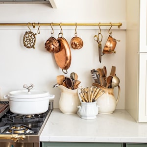 Könnte beinhalten: Eine Küchenszene mit Kupferkochgeschirr, das an einer Messingstange hängt. Ein weißer Topf mit einem Hahngriff steht auf einem Gasherd. Cremefarbene Krüge und ein weißer Behälter halten Küchenutensilien.