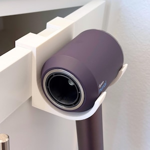 May include: A white plastic wall-mounted hair dryer holder. The holder supports a purple hair dryer with the text "dyson nural". The hair dryer is positioned horizontally, with the nozzle facing forward.