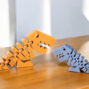 Könnte beinhalten: Zwei dinosaurierförmige Türstopper, einer orange und einer blau, liegen auf einer Holzoberfläche. Der orangefarbene Dinosaurier ist größer und hat ein detaillierteres Skelettdesign. Der blaue Dinosaurier ist kleiner und hat ein einfacheres Skelettdesign.