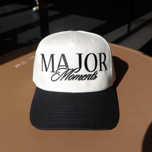 K&ouml;nnte beinhalten: Eine wei&szlig;e Baseballkappe mit schwarzem Schirm. Die Vorderseite der Kappe ist mit schwarzem Stickgarn bestickt und tr&auml;gt den Schriftzug "MAJOR Moments".