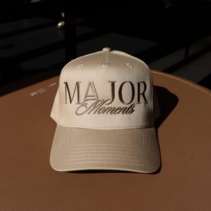 K&ouml;nnte beinhalten: Eine beige Baseballkappe mit brauner Stickerei, auf der "MAJOR Moments" steht.