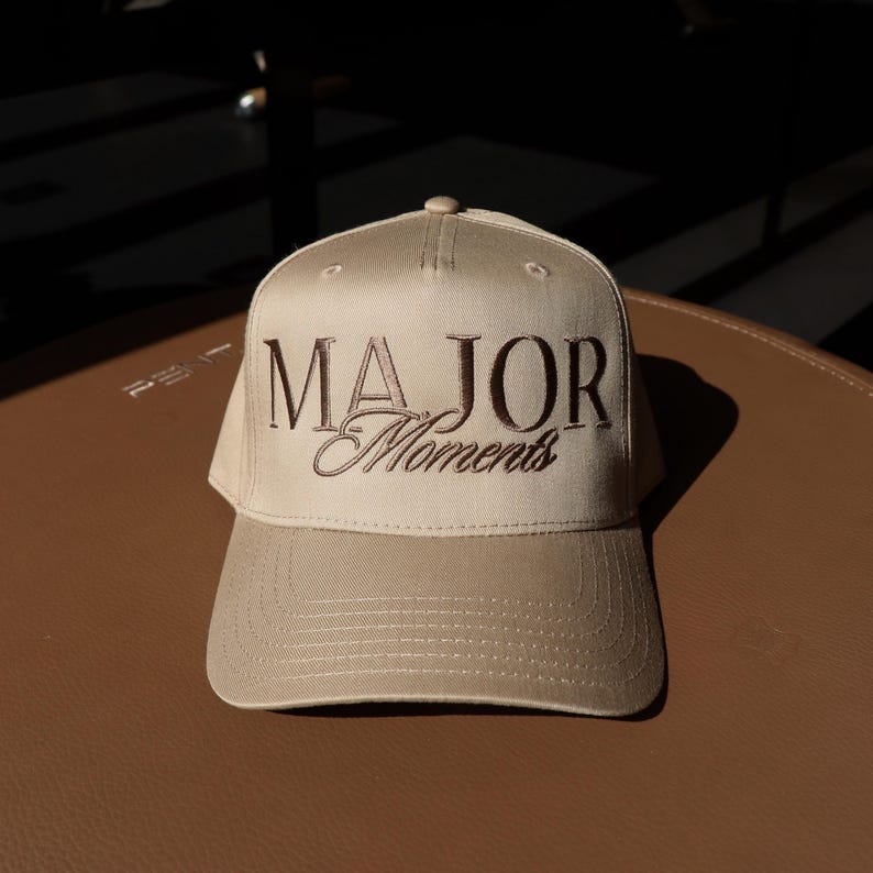 K&ouml;nnte beinhalten: Eine beige Baseballkappe mit brauner Stickerei, auf der "MAJOR Moments" steht.