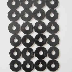 May include: A collection of twenty-four black sewing machine bobbins, each with a central hole and a notched outer edge. The bobbins are arranged in a grid pattern against a white background. Each bobbin has a number printed on it.