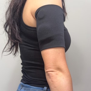 May include: A black sleeveless tank top paired with a matching black arm sleeve that covers the upper arm. The arm sleeve is a solid, plain black fabric that fits snugly around the bicep area, creating a cohesive look with the black tank top.