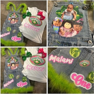 May include: A denim dungaree and white t-shirt set with a Cabbage Patch Kids theme. The dungarees have a green feather trim, patches, and the name "Malani" and the number "One" in pink. The t-shirt has a pink bow.