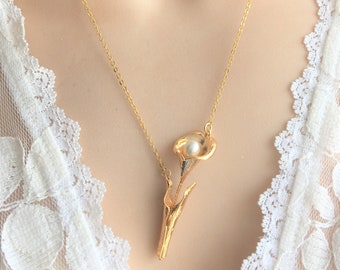 Puede incluir: Un collar dorado con un colgante de flor de cala con una perla en el centro. El colgante tiene detalles plateados y dorados y cuelga de una fina cadena dorada. El collar se muestra sobre un fondo de encaje blanco.