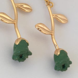 May include: A pair of gold-coloured earrings featuring a rose design. Each earring has a gold stem with two leaves and a green rosebud at the bottom. The earrings have a hook closure.