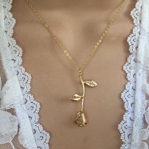May include: A gold rose pendant necklace on a model wearing a white lace top.