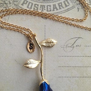 May include: Gold chain necklace with a blue rose charm and a small gold charm with the letter 'S' on it. The necklace is on a vintage postcard with the word 'POSTCARD' printed on it.