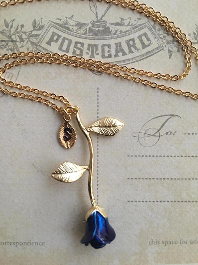 May include: Gold chain necklace with a blue rose charm and a small gold charm with the letter 'S' on it. The necklace is on a vintage postcard with the word 'POSTCARD' printed on it.