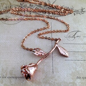 May include: A rose gold necklace with a delicate rose pendant. The rose has a long stem and two leaves. The chain is a simple, linked chain.