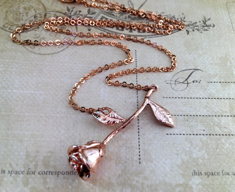 May include: A rose gold necklace with a delicate rose pendant. The rose has a long stem and two leaves. The chain is a simple, linked chain.