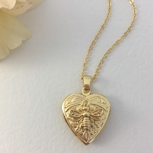 May include: A gold heart-shaped locket necklace featuring a bee design. The locket has a detailed floral pattern and a 3D bee in the centre.