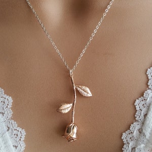 May include: A rose gold necklace with a delicate rose pendant. The rose is made of metal and has a textured finish. The necklace is on a model wearing a white lace top.