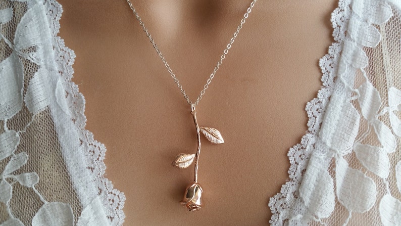 May include: A rose gold necklace with a delicate rose pendant. The rose is made of metal and has a textured finish. The necklace is on a model wearing a white lace top.
