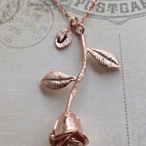 May include: A rose gold necklace with a rose pendant. The rose is detailed with leaves and a stem. The necklace is on a white background.