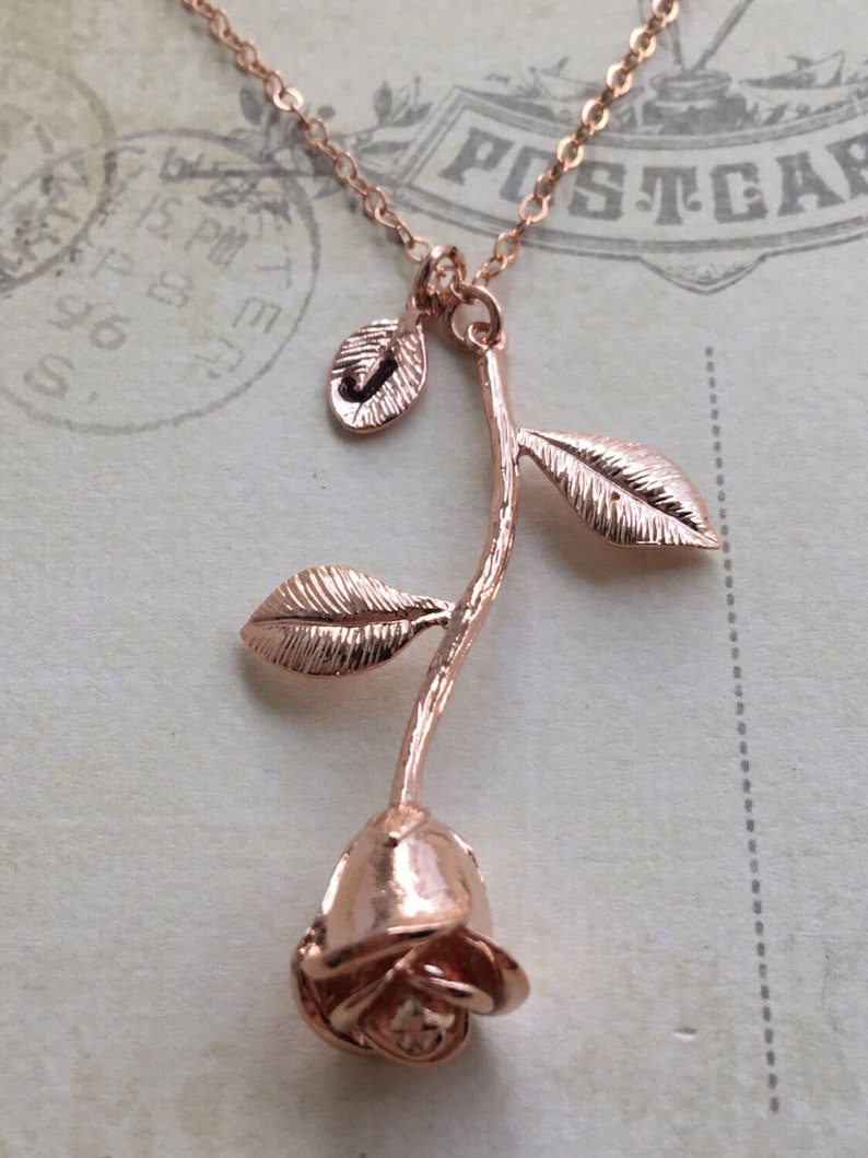 May include: A rose gold necklace with a rose pendant. The rose is detailed with leaves and a stem. The necklace is on a white background.