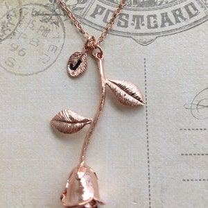 May include: Rose gold necklace with a single rose pendant and a small leaf charm with the letter J. The necklace is on a vintage postcard.