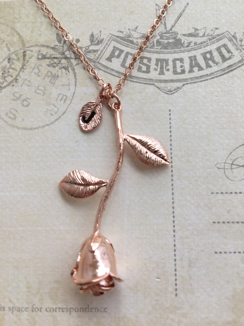 May include: Rose gold necklace with a single rose pendant and a small leaf charm with the letter J. The necklace is on a vintage postcard.