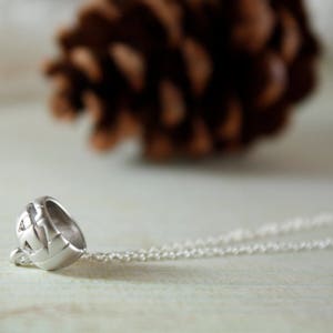 May include: A silver necklace with a detailed pendant shaped like a small, round helmet. The necklace is lying on a white surface.