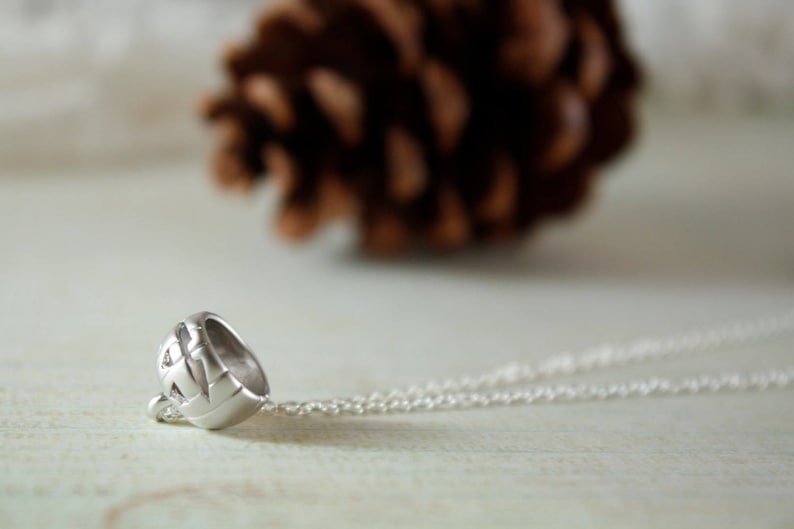 May include: A silver necklace with a detailed pendant shaped like a small, round helmet. The necklace is lying on a white surface.