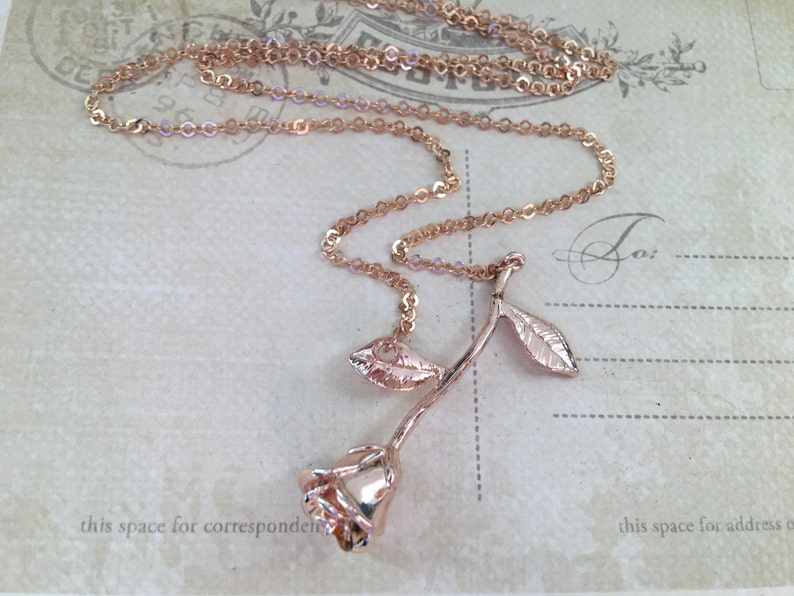 May include: A rose gold necklace with a rose pendant. The rose has a long stem and two leaves. The chain is a delicate, cable chain.